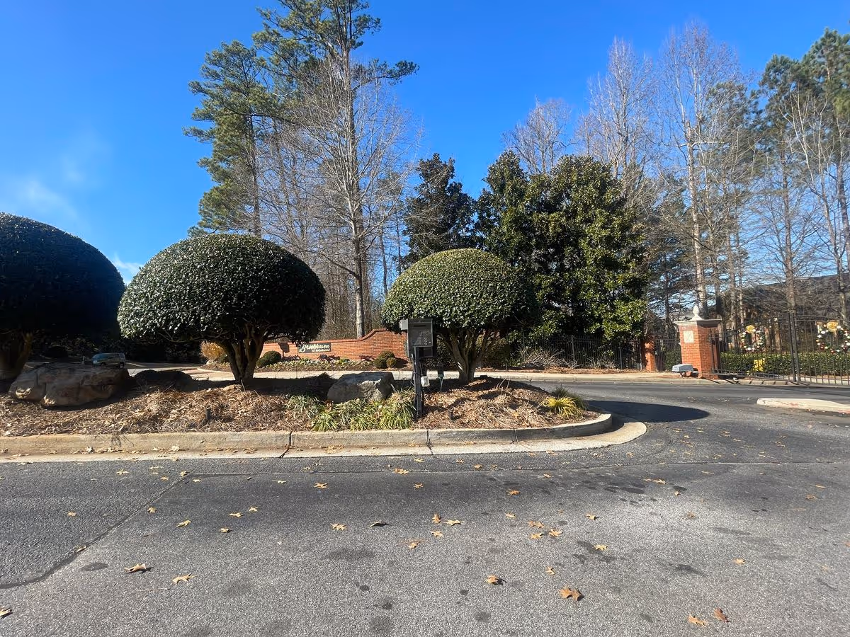 Entrance driveway and landscaped island with trimmed round shrubs and brick entry columns under a clear blue sky.