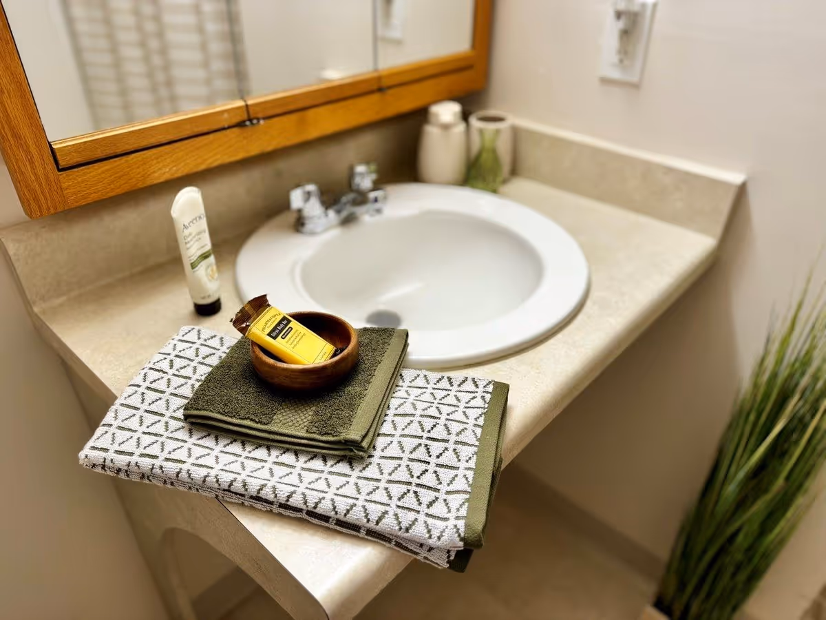 Bathroom vanity with a round sink, folded towels, a wooden bowl with soap, and toiletries under a wooden-framed mirror.