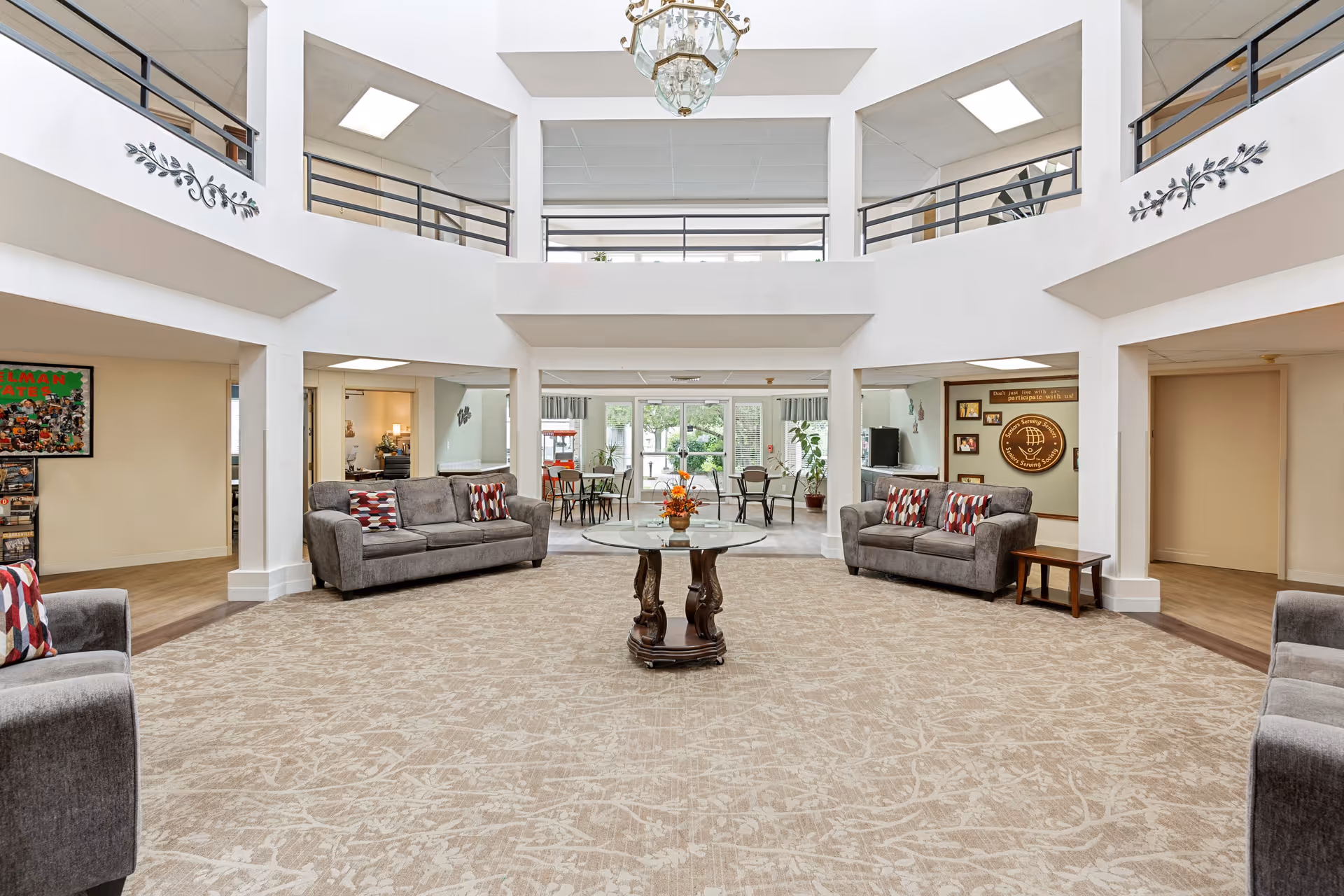 Spacious and well-lit common area in a senior living facility with beige patterned carpet, two gray sofas with patterned cushions, a round glass-top table with a floral centerpiece in the center, and a chandelier hanging from the ceiling. The area has a high ceiling with a second-floor balcony railing visible, and large windows in the background letting in natural light. There are also some chairs and tables near the windows and decorative wall accents.