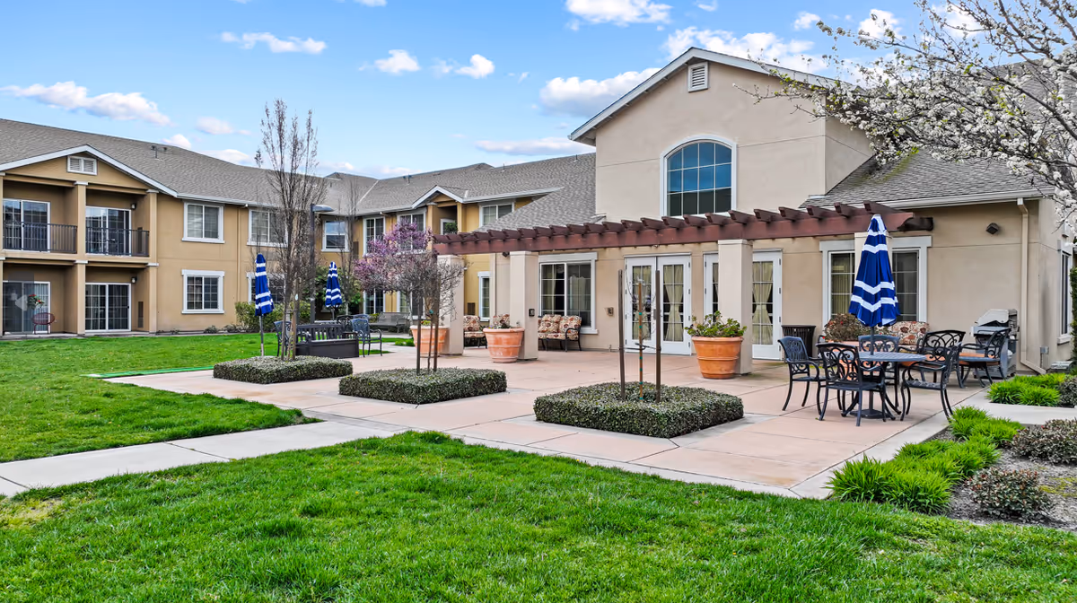 Outdoor patio area of Manteca Assisted Living and Memory Care facility with tables, chairs, blue and white striped umbrellas, potted plants, and a pergola attached to the building. The building has beige walls and multiple windows, surrounded by green grass and landscaped bushes under a partly cloudy sky.