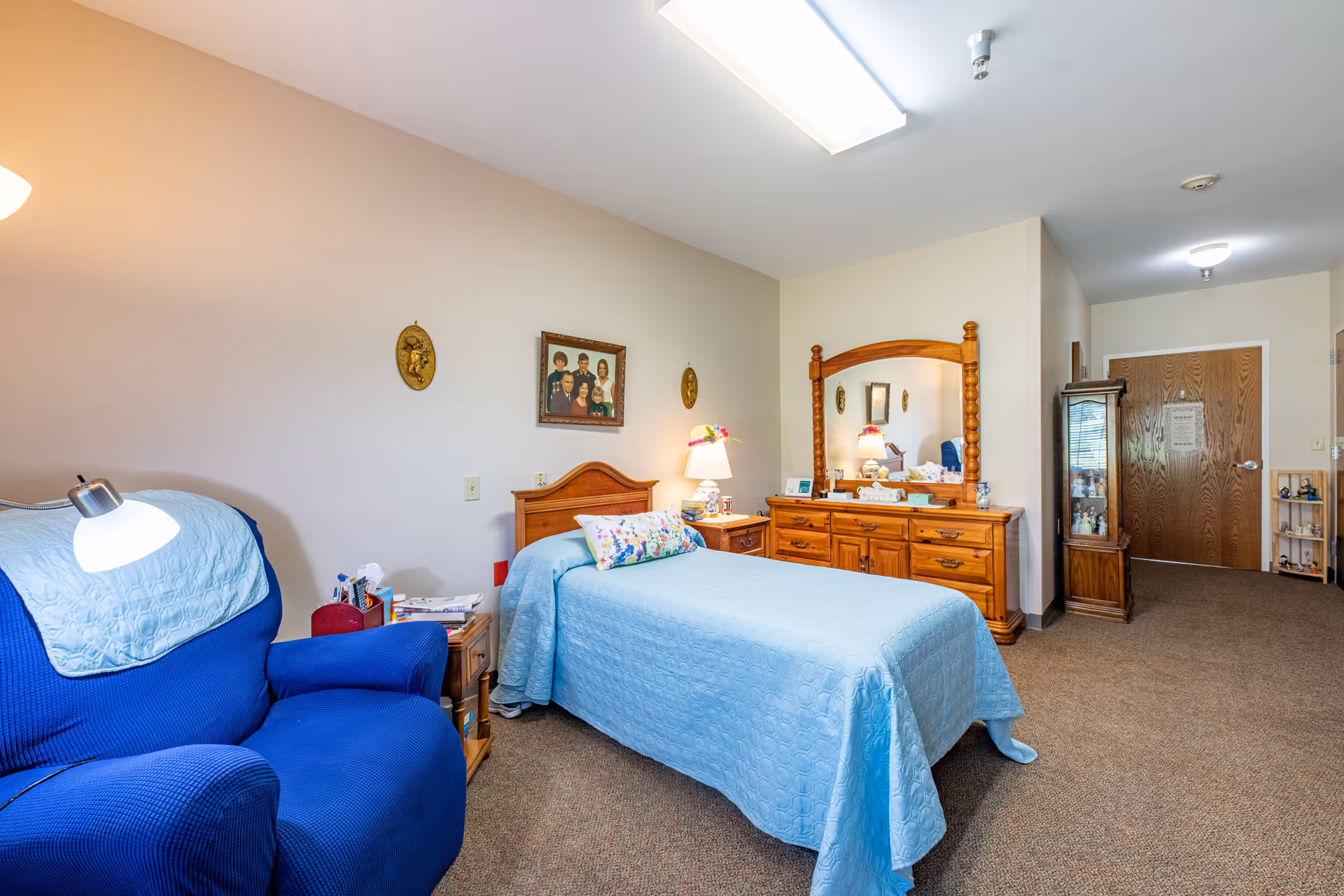 A cozy bedroom in an assisted living facility featuring a single bed with a light blue quilt and floral pillow, a wooden dresser with a large mirror, a blue armchair with a matching light blue cover, and family photos and decorative wall hangings on the beige walls.