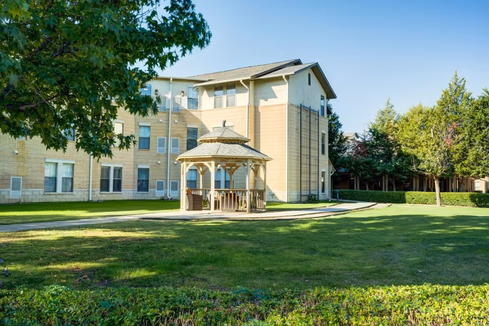 Outdoor view of a senior living facility with a beige multi-story building in the background, a wooden gazebo with seating in the center, surrounded by green grass, trees, and shrubs under a clear blue sky.