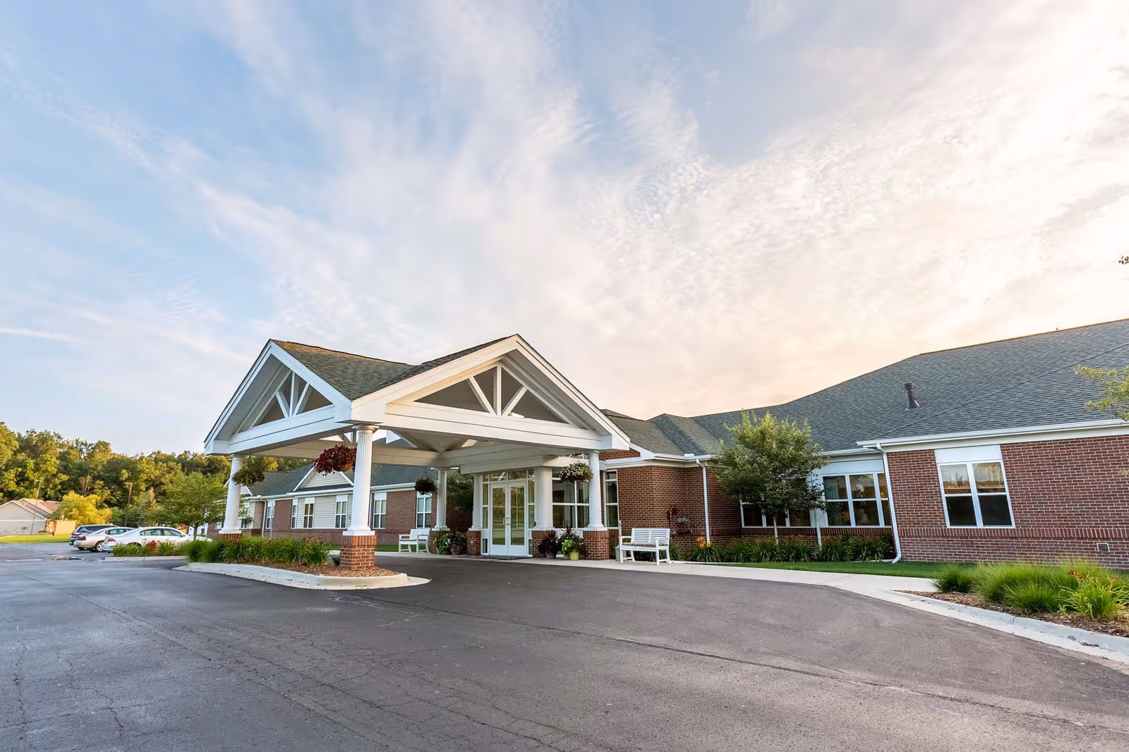 Covered entrance of a single-story brick senior living building with a porte-cochere, benches, and a paved driveway.