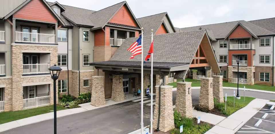 Exterior view of a senior living facility named Harmony At Bellevue showing a large covered entrance with stone pillars, two flagpoles with American and state flags, and a multi-story building with balconies and windows under a cloudy sky.