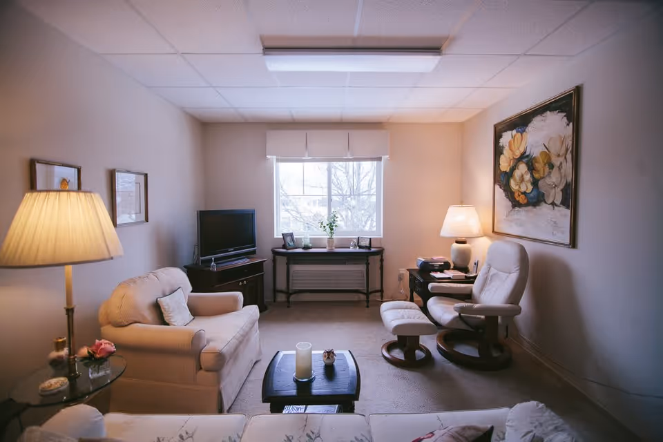 A cozy living room with a beige armchair, a white recliner with matching ottoman, a small dark wooden coffee table with candles, a TV on a stand, a window with a white valance, and a large floral painting on the wall. The room is softly lit by two lamps and has a carpeted floor.
