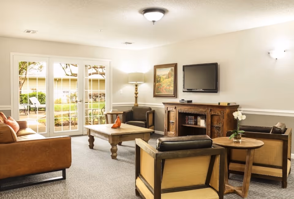 A cozy living room with a brown leather sofa, two armchairs with beige cushions, a wooden coffee table with a decorative item, a wooden side table with a vase of white flowers, a floor lamp, a wall-mounted flat-screen TV, and a framed painting. French doors open to an outdoor garden area.