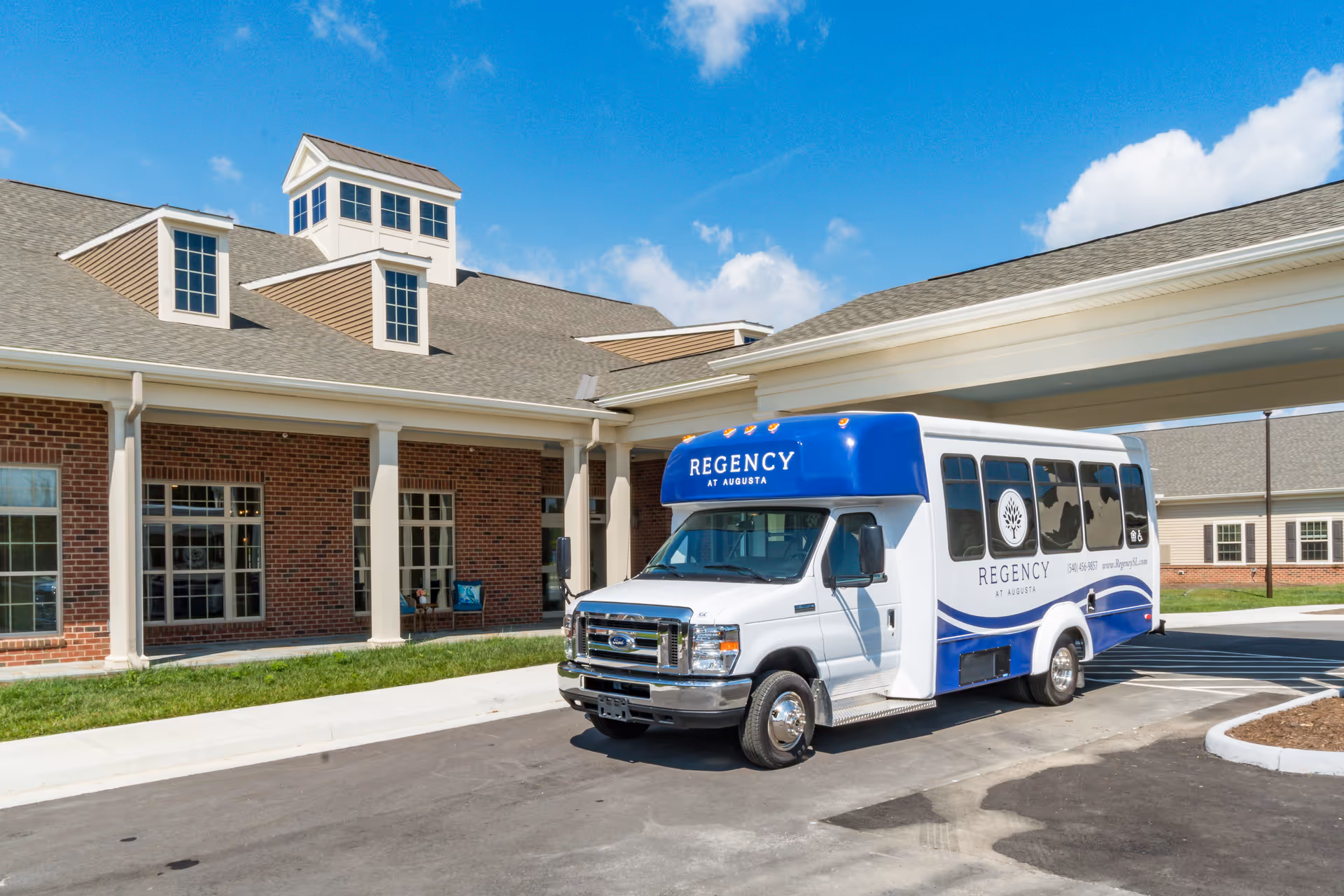 White and blue Regency at Augusta shuttle parked under the covered entrance of a brick senior living building.
