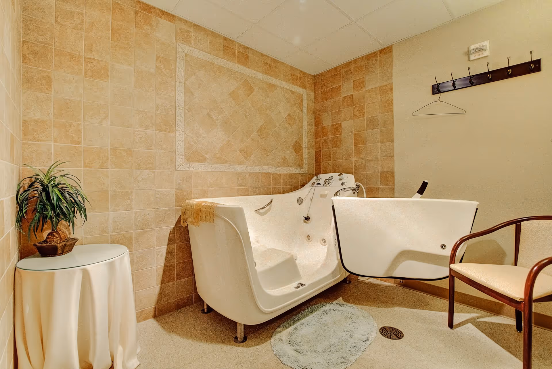 A beige tiled bathroom with a walk-in bathtub featuring a door for easy access. There is a small round table with a potted plant on it, a wooden chair with a cushioned seat, a wall-mounted coat rack with a single hanger, and a small round bath mat on the floor.