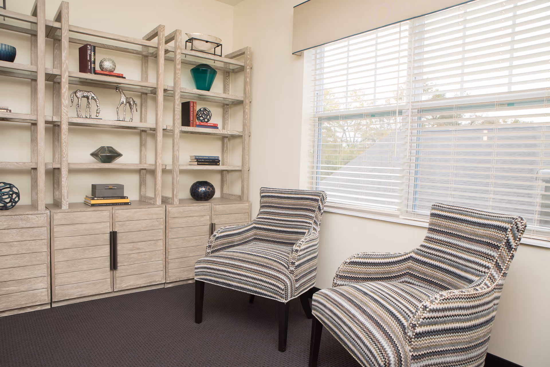 A cozy interior room with two patterned armchairs placed near a window with white blinds. Behind the chairs is a light wood shelving unit with various decorative items including books, sculptures, and vases.