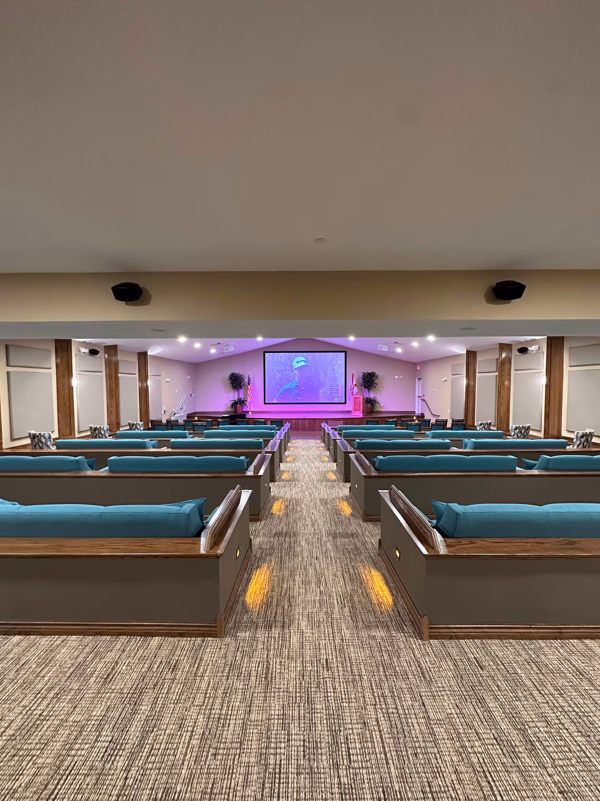 Interior view of a large room with rows of cushioned benches with teal upholstery facing a stage with a large screen displaying a person wearing a cap. The room has carpeted floors, beige walls with wooden trim, and soft lighting along the aisle.