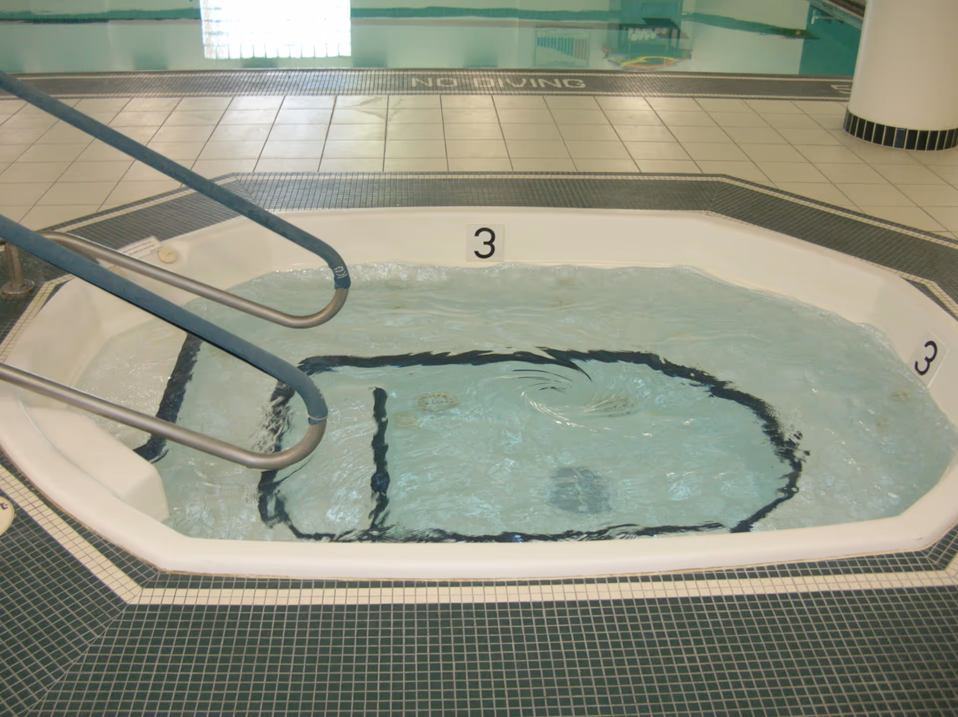 Indoor hot tub/jacuzzi with metal handrails and tiled deck next to a swimming pool.