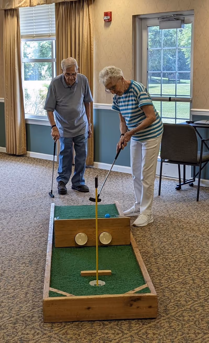 Two elderly individuals playing indoor mini golf in a carpeted room with large windows and a glass door. One person is putting while the other watches, both holding putters. The room has beige walls with a chair and table in the background.