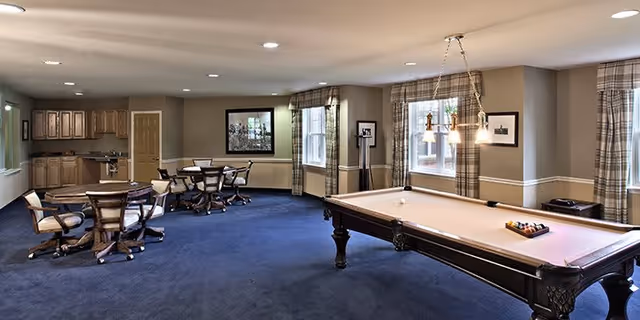 Spacious interior community room with a pool table, several card tables and chairs, and a kitchenette under recessed lighting.