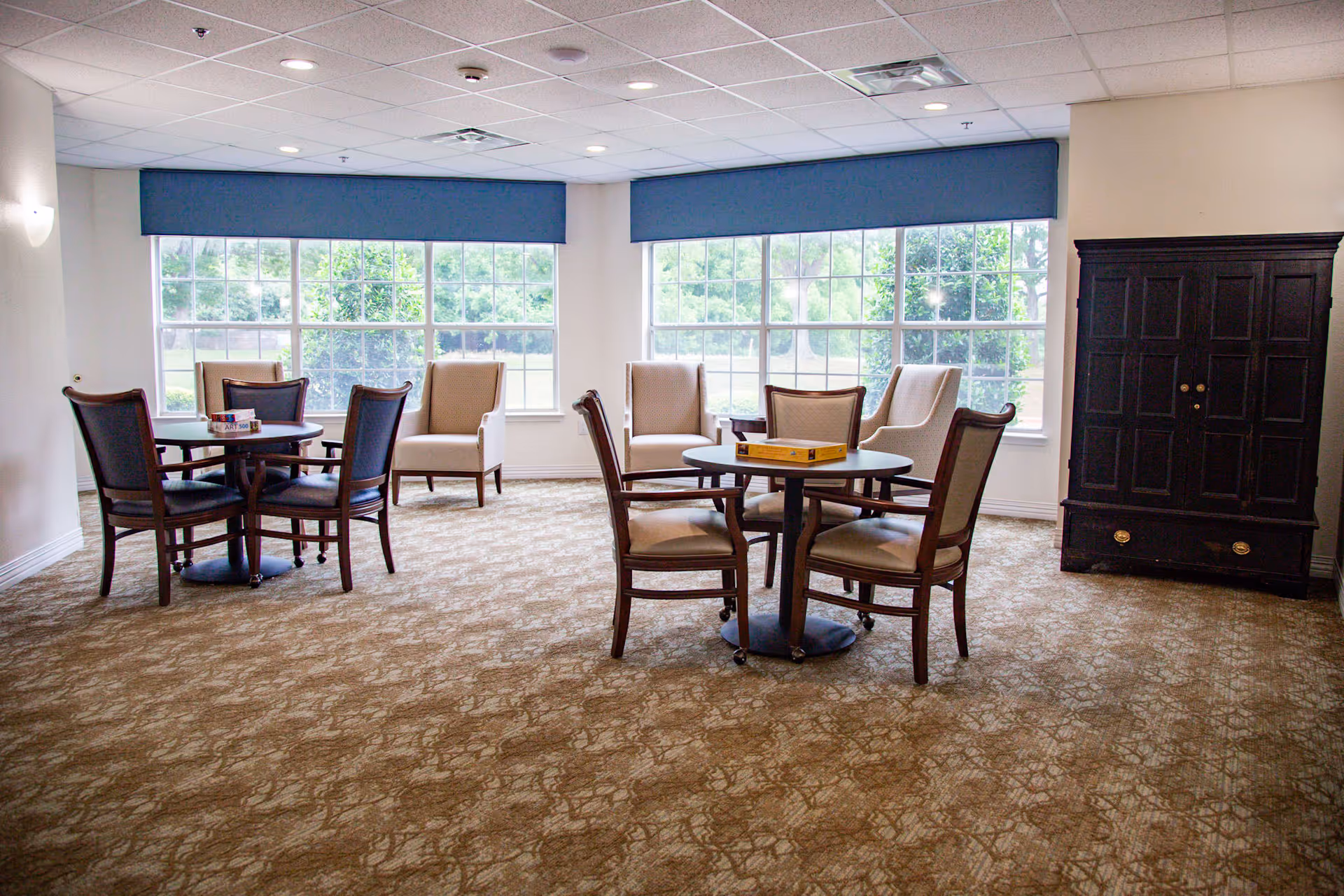 A bright room with large windows covered by blue valances, featuring two round tables each surrounded by four chairs. There are additional beige armchairs placed near the windows and a dark wooden cabinet against the wall on the right side.