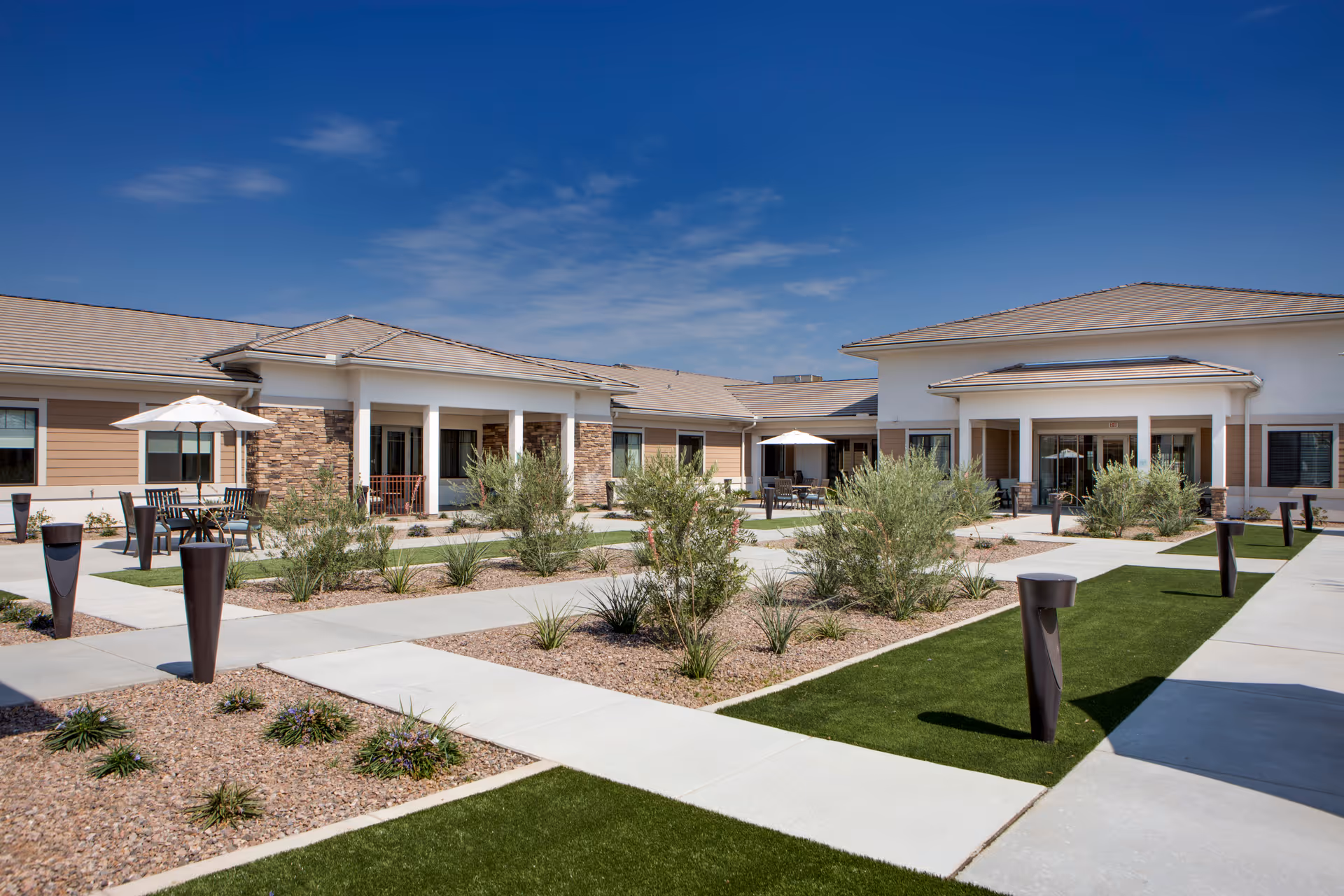 Sunlit courtyard with paved walkways, landscaping, outdoor seating and single-story residential buildings under a blue sky.