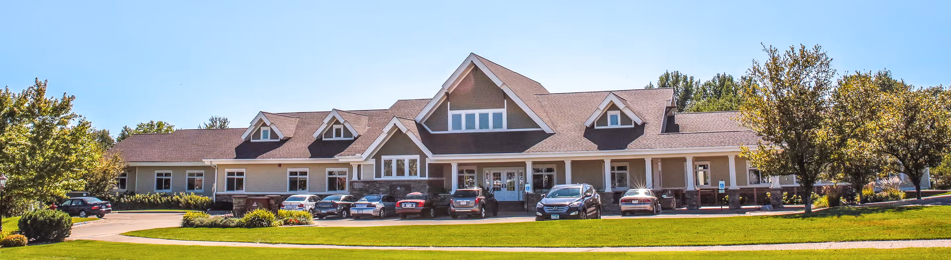 Front exterior view of a large, single-story assisted living facility building with a peaked roof and multiple dormer windows. Several cars are parked in front of the building, which is surrounded by green lawns and trees under a clear blue sky.