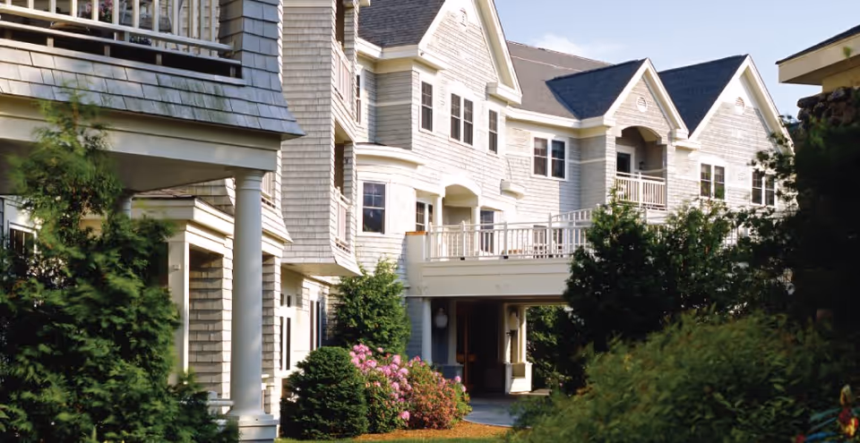 Exterior front view of a multi-story shingle-sided senior living building with balconies and landscaped shrubs.