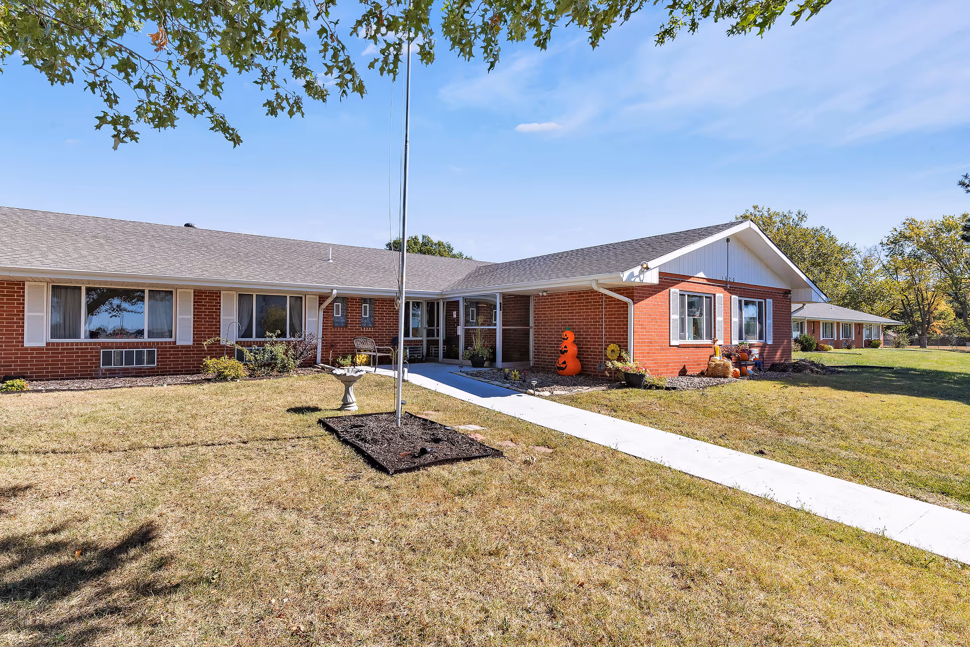 Exterior view of a single-story brick building with white trim, surrounded by a grassy lawn and trees under a clear blue sky. A concrete walkway leads to the entrance, which is decorated with autumn-themed items including a pumpkin figure and potted plants.
