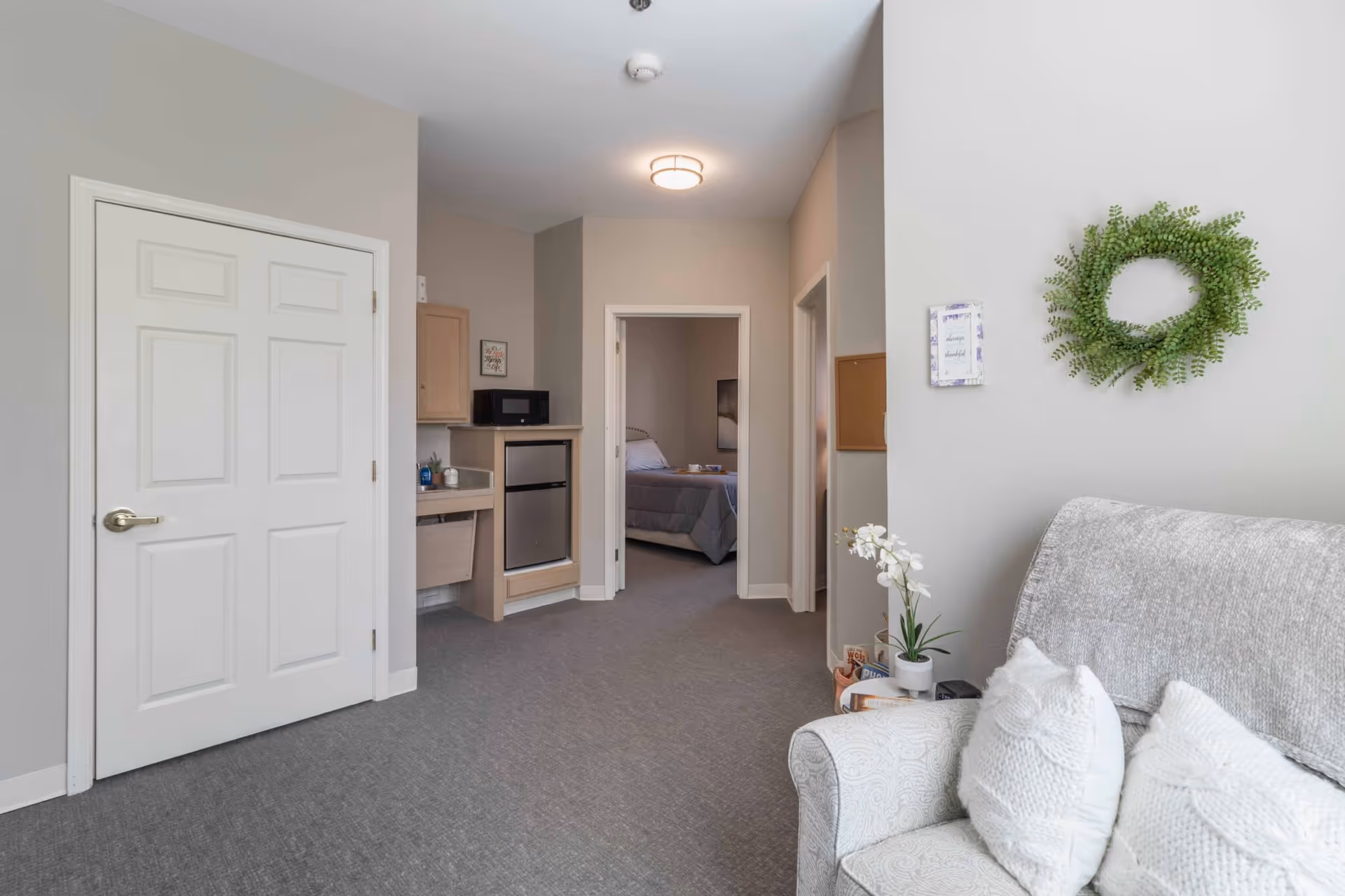 A cozy interior room with a gray armchair adorned with white pillows, a small side table with a potted orchid, and a green wreath hanging on the wall. In the background, there is a small kitchenette area with a microwave and mini fridge, and a doorway leading to a bedroom with a made bed.