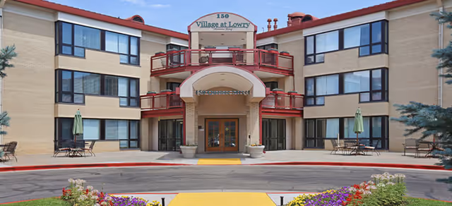 Front exterior view of a three-story senior living facility named Village at Lowry with beige brick walls, large windows, red balcony railings, and a main entrance with double glass doors. There are outdoor seating areas with tables and umbrellas on either side of the entrance, and colorful flowers planted in front of the building.