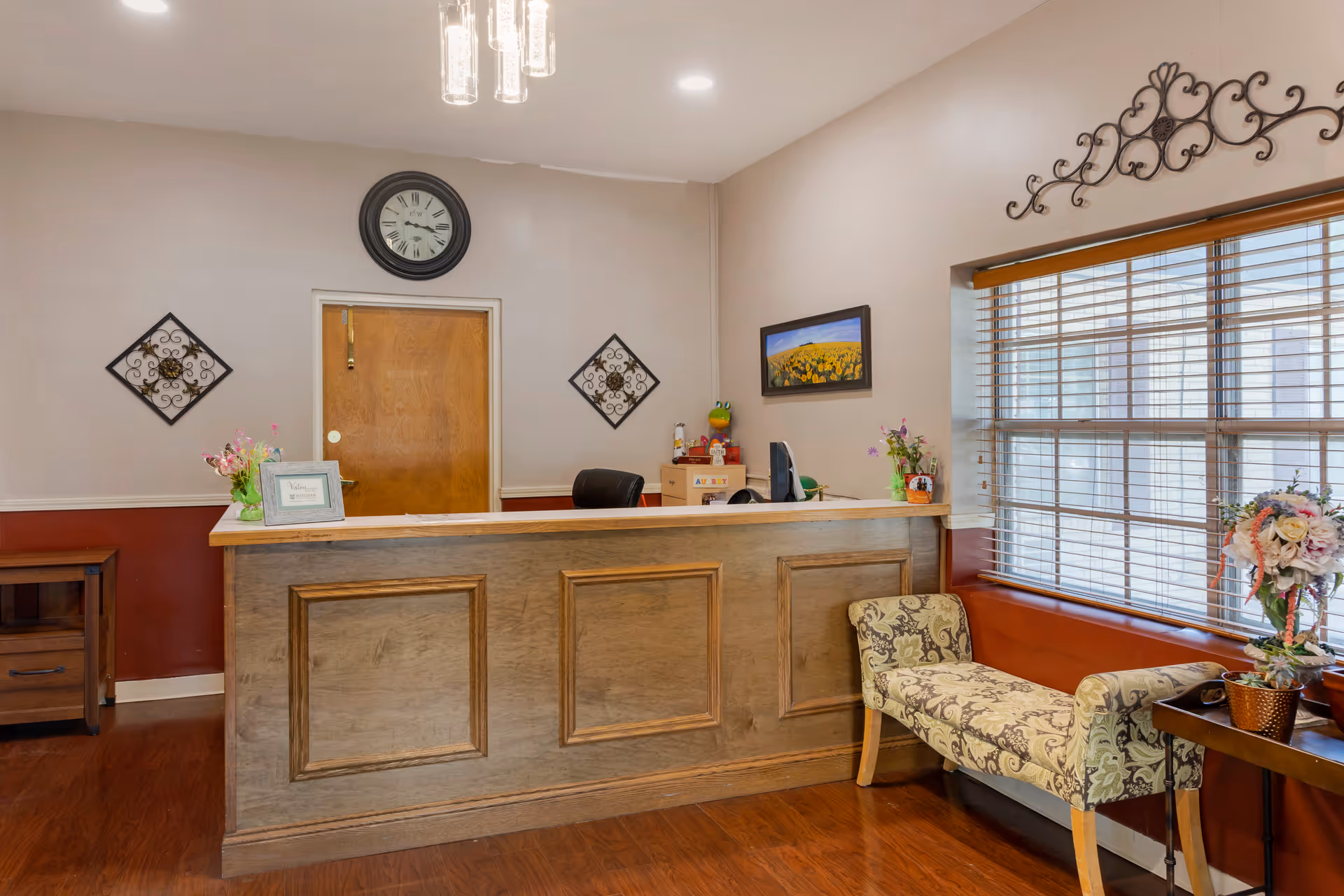 Reception area with a wooden front desk, a clock on the wall above a wooden door, decorative wall hangings, a patterned bench near a window with blinds, and flower arrangements on the desk and a side table.
