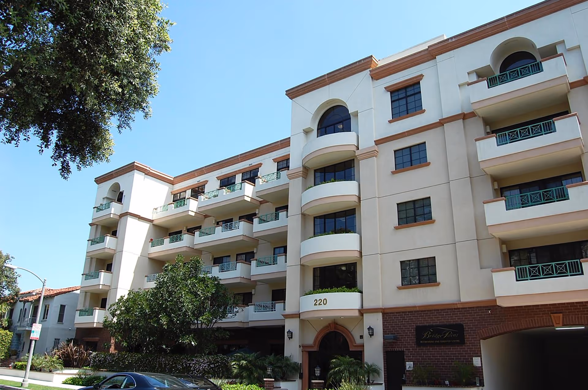 Exterior view of a multi-story residential building with balconies, greenery, and a clear blue sky.
