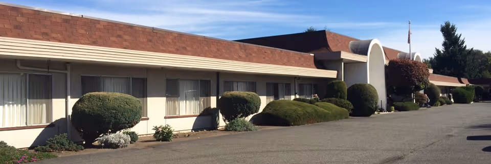 Exterior view of a single-story building with a brown roof and beige walls, featuring several windows with curtains and neatly trimmed bushes along the front. The sky is clear and blue.