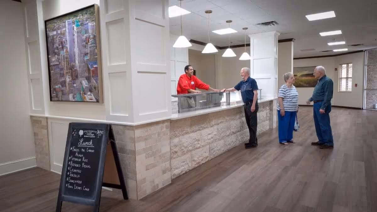 Staff member behind a serving counter talks with three elderly residents in a spacious retirement community dining area.