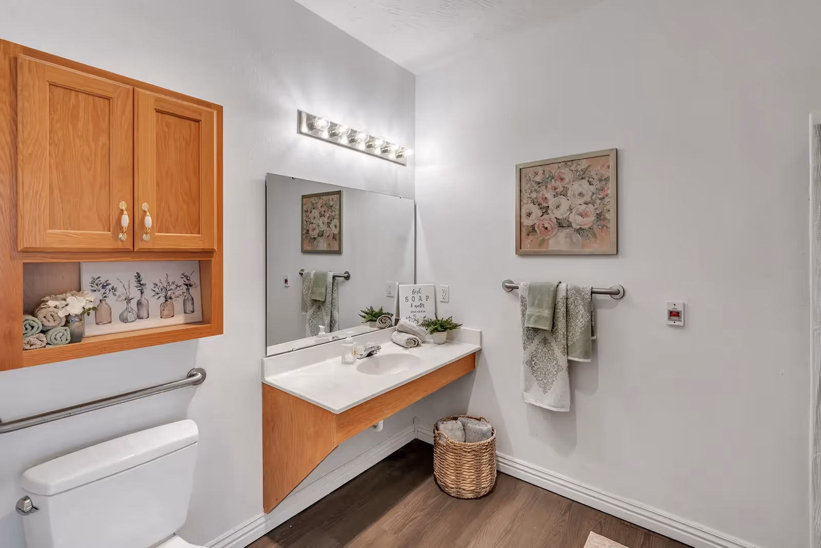 Well-lit bathroom with a wall-mounted sink and mirror, wooden storage cabinet, toilet, towel racks, and decorative accents.