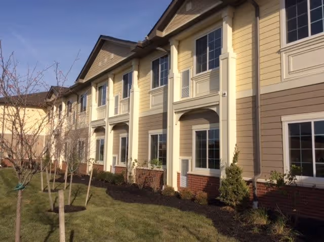Exterior view of a multi-story residential building with beige and brown siding, white trim, and multiple windows. There is a landscaped area with small trees and shrubs in front of the building under a clear blue sky.