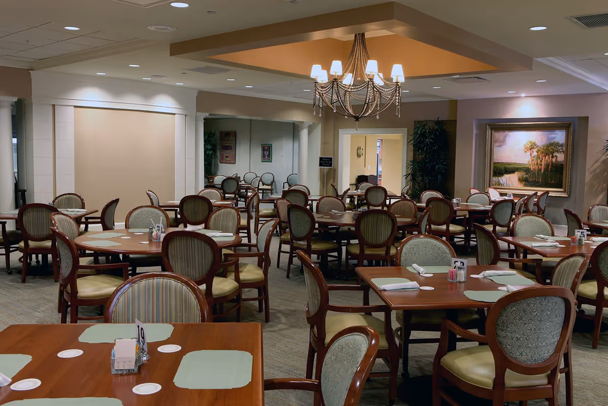 A spacious dining room with multiple wooden tables and cushioned chairs arranged neatly. Each table has placemats, napkins, and small condiment holders. A decorative chandelier hangs from the ceiling, and a large framed painting of a landscape with palm trees is on the wall. The room has soft lighting and a warm, inviting atmosphere.