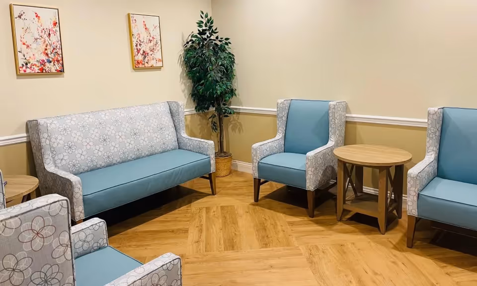 A cozy seating area with a light blue patterned sofa and matching armchairs arranged around a wooden side table. The room has light beige walls with white trim, two floral paintings on the wall, and a potted plant in the corner. The floor is a warm wood tone.