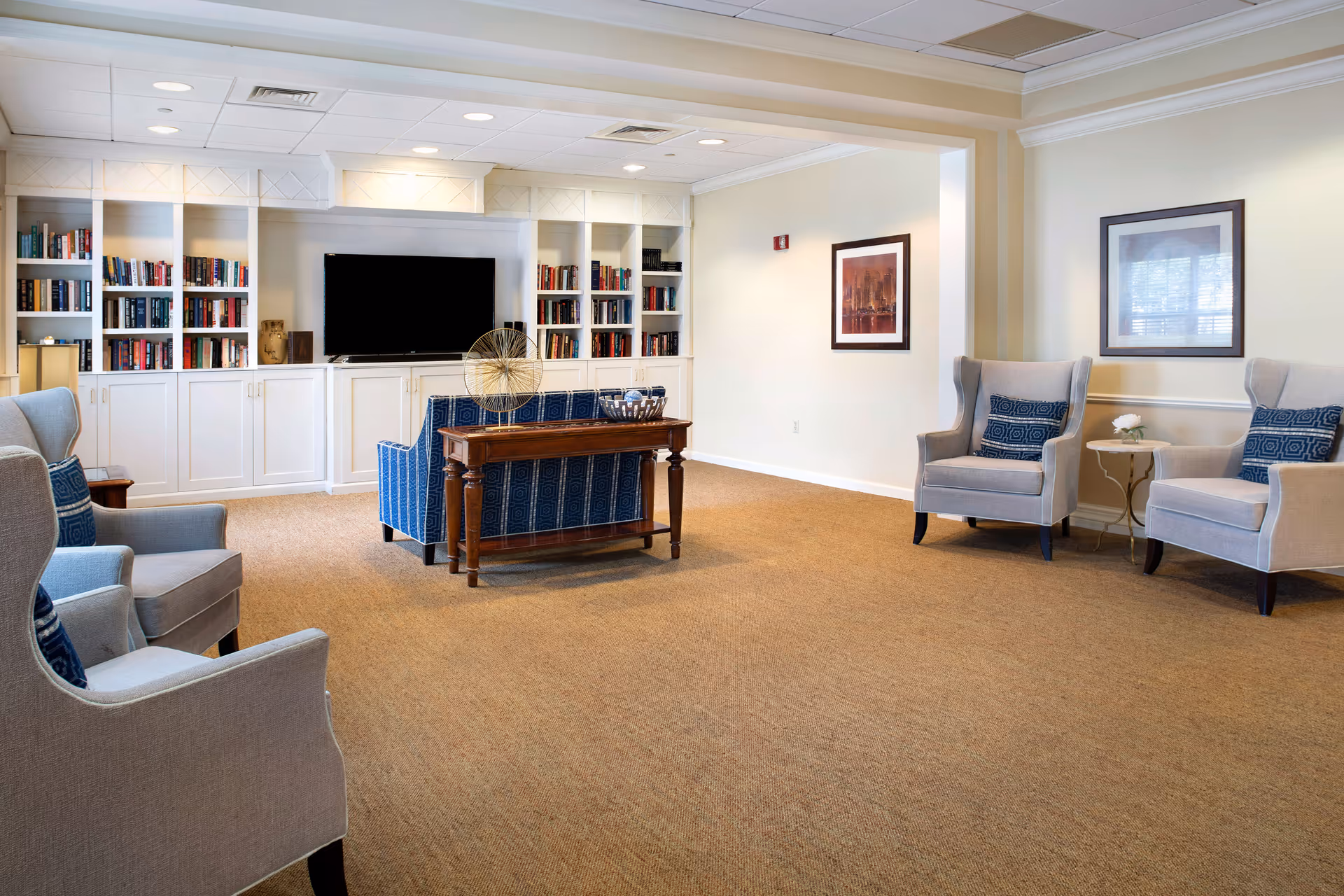 Bright carpeted common lounge with armchairs, a sofa, built-in bookshelves and a television.