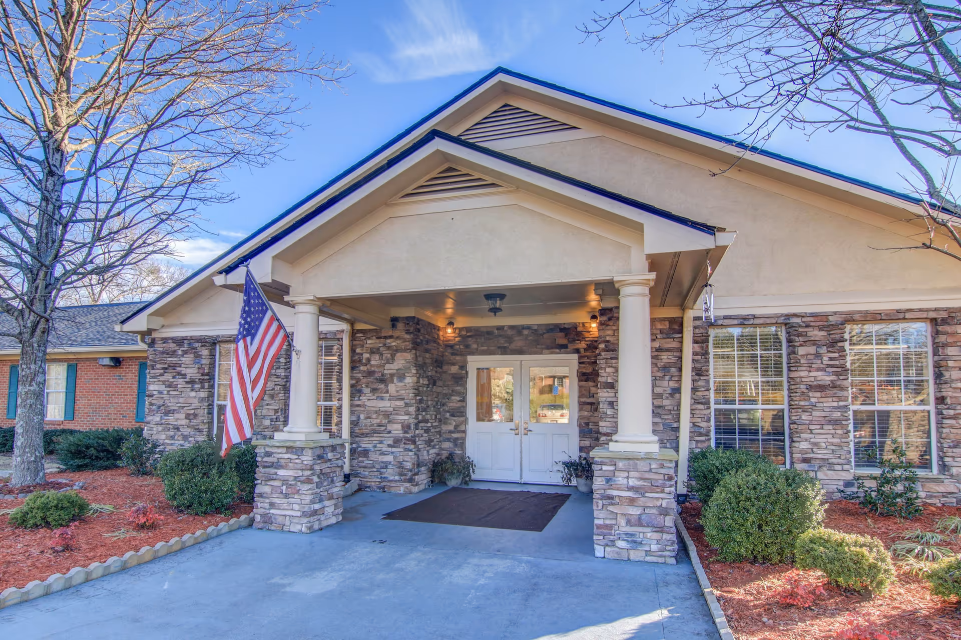 Front entrance of Manchester Court Assisted Living and Memory Care facility featuring a covered porch with stone pillars, an American flag mounted on the left pillar, double white doors, and landscaped bushes and trees with mulch beds on either side.