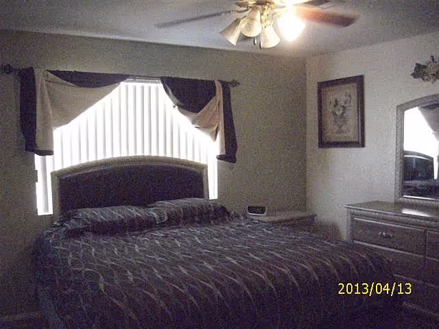 Bedroom with a large bed beneath an arched window with vertical blinds, a ceiling fan, dresser with mirror, and framed wall art.