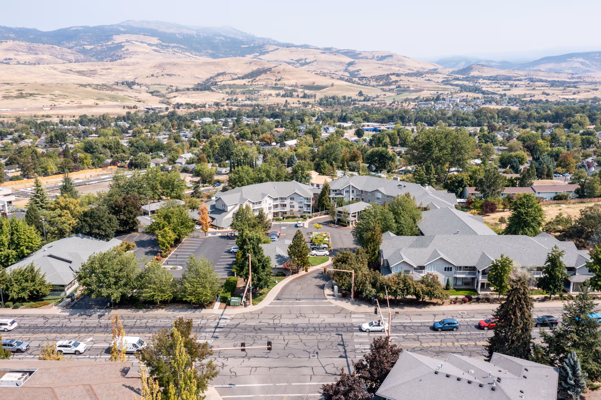 Aerial view of Maple Ridge Senior Living facility surrounded by trees and residential areas, with mountains in the background and a main road with cars in the foreground.