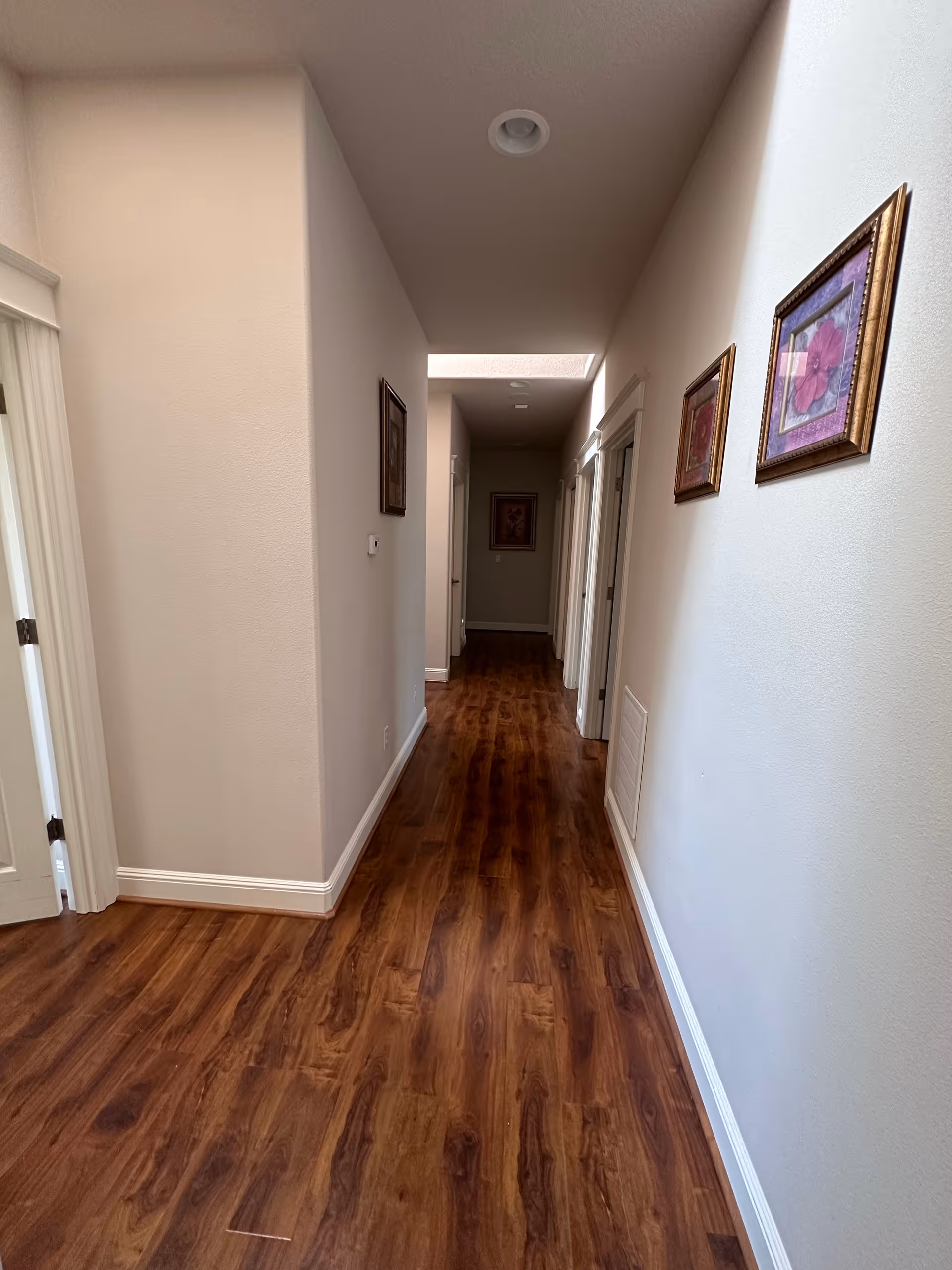 A long indoor hallway with wooden flooring and white walls. Several framed pictures hang on the right wall, and there are multiple doorways along the hallway. The ceiling has recessed lighting and a skylight near the end of the hallway.