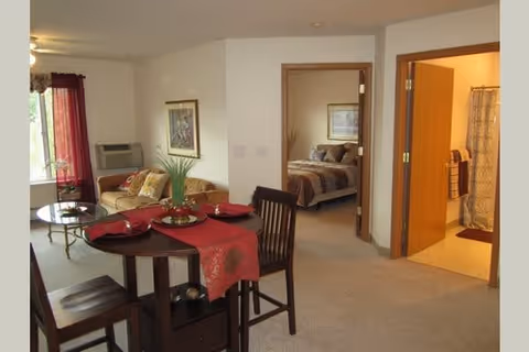 Interior view of a furnished senior living apartment with a dining table in the foreground, a living area to the left, and a bedroom and bathroom visible through open doorways.