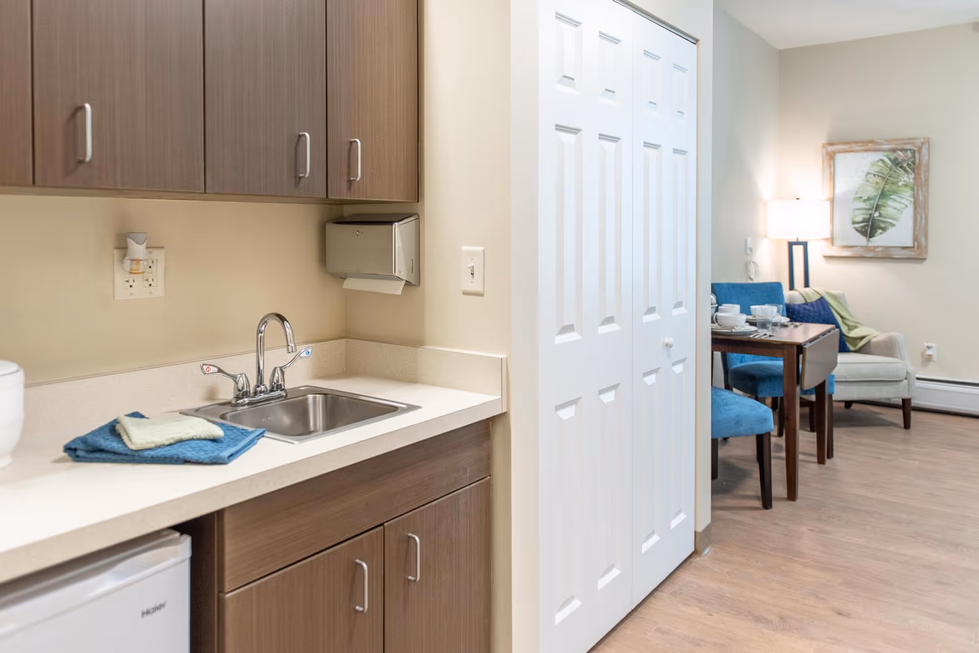 Small kitchenette with a sink and wood cabinets opening to a dining and seating area with blue chairs and wall art.