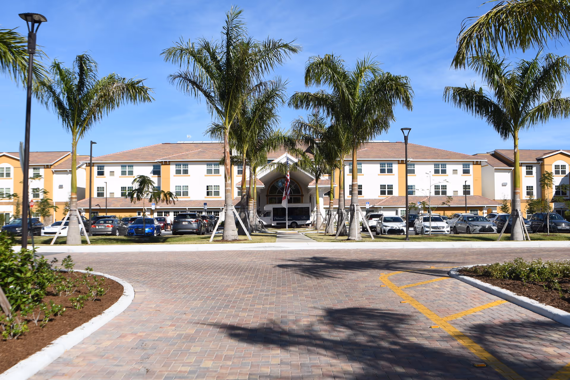 Front exterior view of Palomino Gardens Retirement Community building with a paved driveway, palm trees, and parked cars under a clear blue sky.