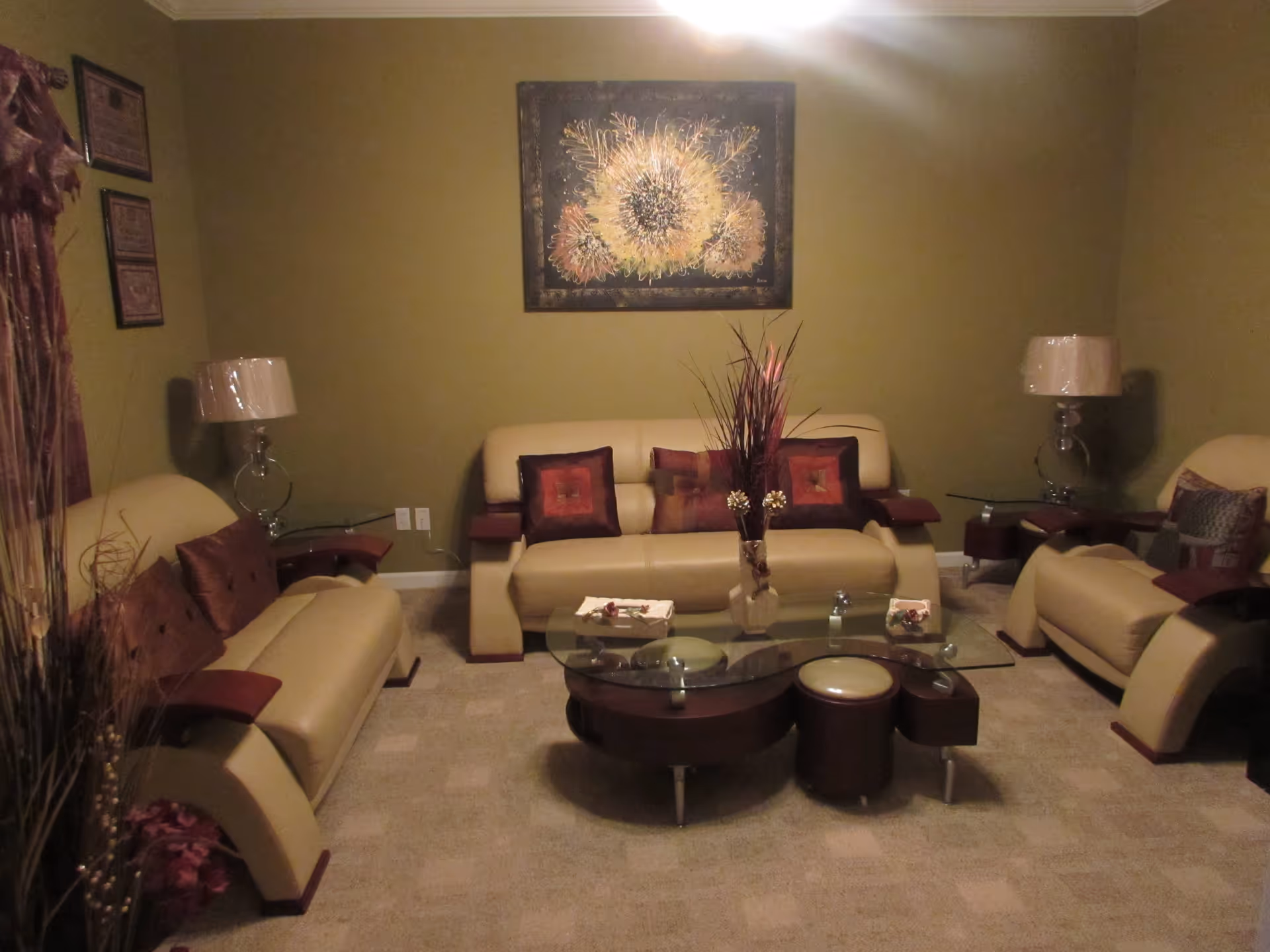 A cozy living room with beige leather sofas and armchairs arranged around a glass coffee table. The walls are painted olive green, and a floral painting hangs above the central sofa. Two lamps with beige shades are placed on side tables beside the sofas. Decorative pillows and a vase with tall dried plants are on the coffee table.