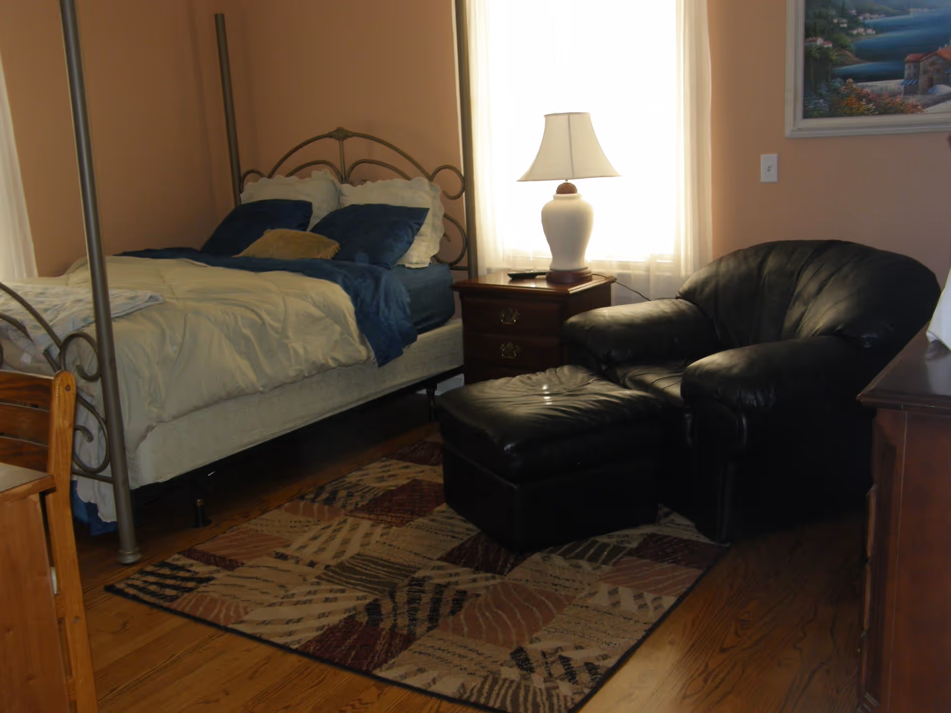 A cozy bedroom with a metal frame bed made up with white and blue bedding, a wooden nightstand with a white lamp, a black leather armchair with matching ottoman, a patterned area rug on a wooden floor, and a framed painting on the wall.
