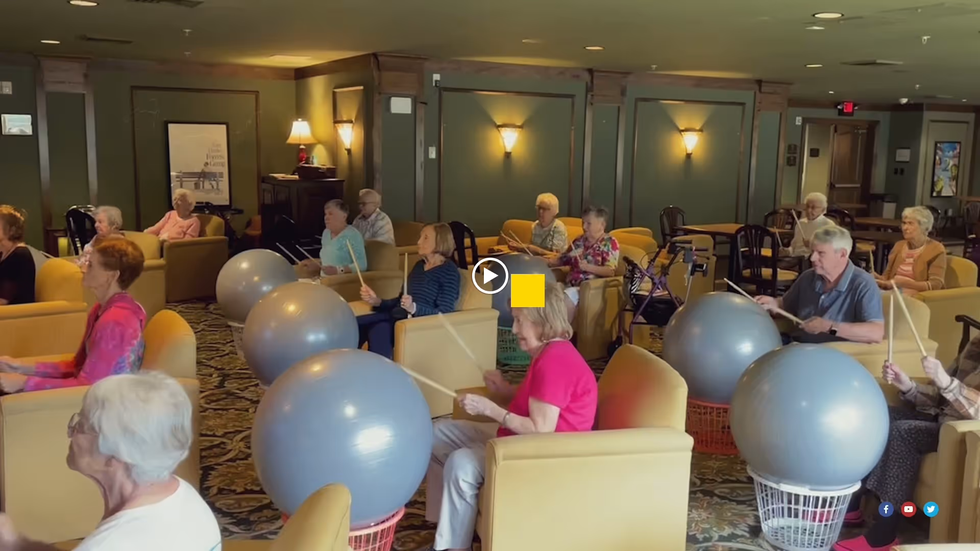 A group of seniors seated in a communal lounge using large exercise balls and drumsticks for a group activity.