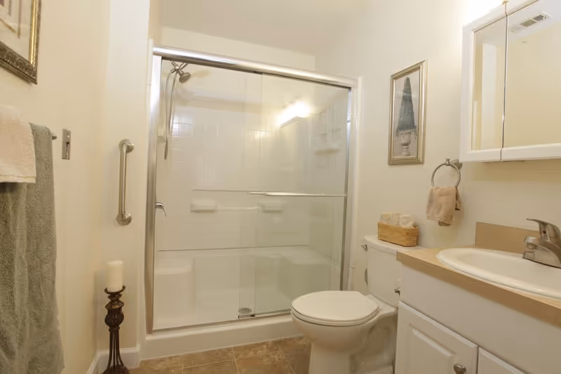 A clean bathroom featuring a glass-enclosed shower with a built-in seat, a white toilet, a vanity with a sink and faucet, a wall-mounted mirror cabinet, a towel ring with a beige towel, a framed picture on the wall, and a decorative candle holder on the floor. Towels hang on a rack on the left wall.
