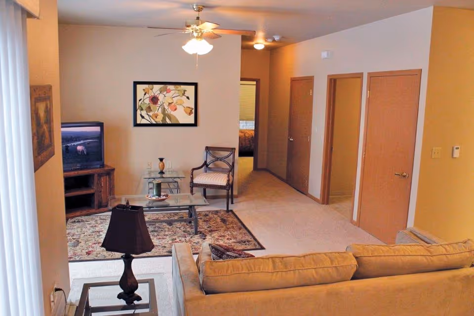 Living room with a sofa, glass coffee table, armchair, TV on a stand, ceiling fan, and a hallway with multiple doors.