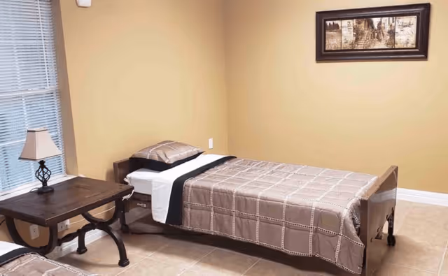 A simple bedroom with a single bed covered in a beige and brown checkered bedspread, a matching pillow, a wooden side table with a small lamp, beige walls, and a framed picture hanging above the bed.