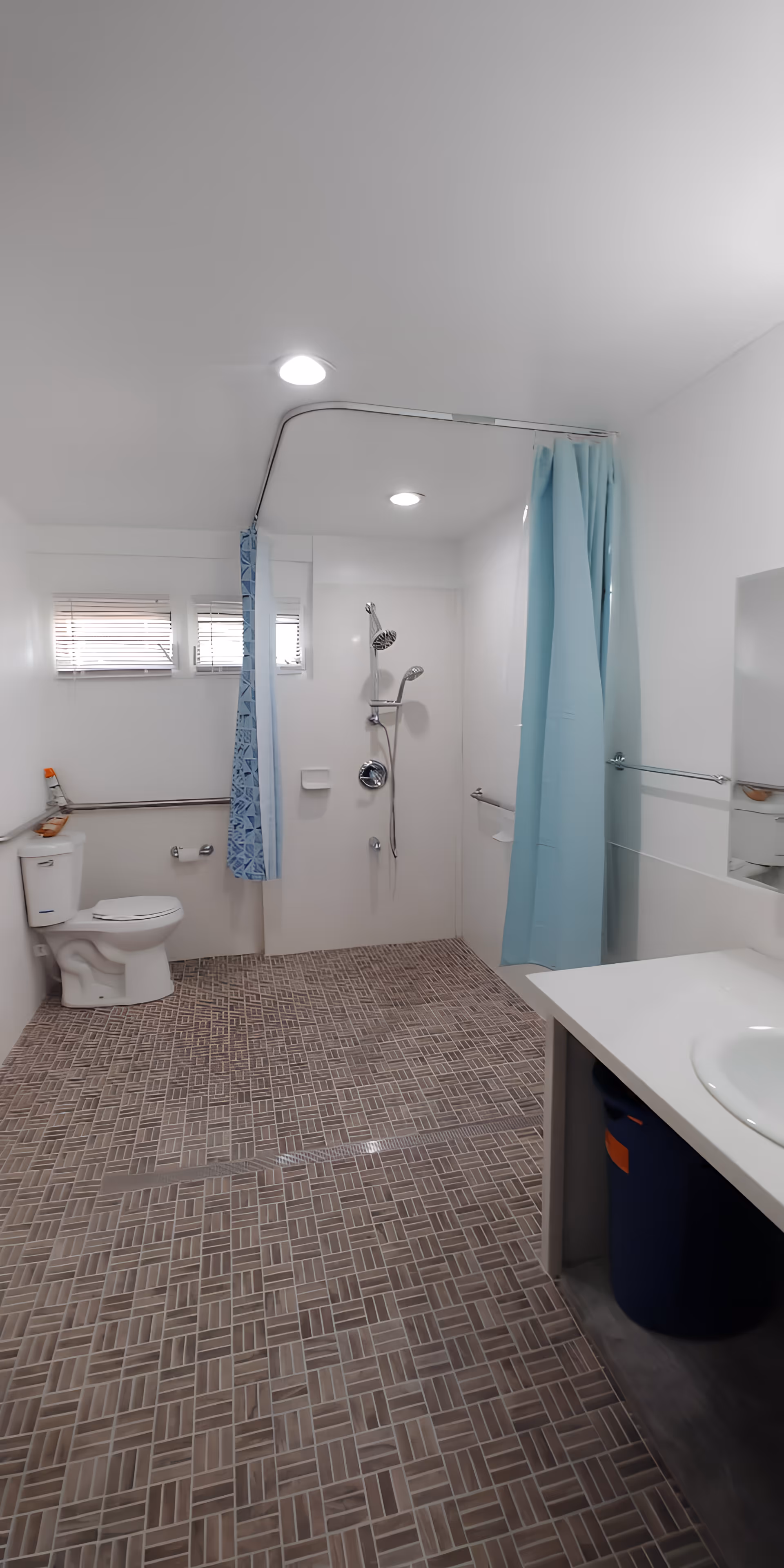 A clean, accessible bathroom with a toilet, a walk-in shower area with two shower heads and a blue shower curtain, a sink with a countertop, and grab bars installed near the toilet and shower area. The floor has a patterned tile design, and there are small windows near the ceiling allowing natural light in.