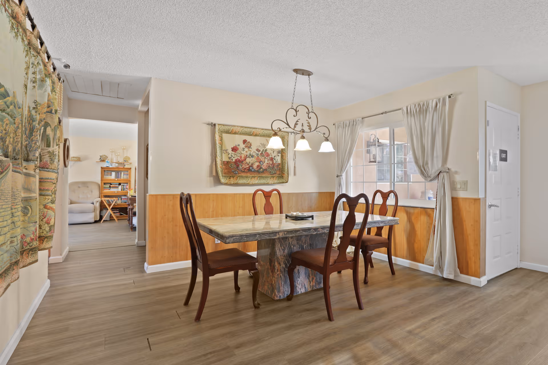 A dining room with a rectangular marble table and four wooden chairs with cushioned seats. A decorative hanging light fixture with three lamps is above the table. The walls are painted beige with wooden paneling on the lower half. There is a window with cream curtains tied back, and a door labeled 'EXIT' on the right side. A floral tapestry hangs on the wall above the paneling. In the background, a living area with a beige armchair and a wooden bookshelf is partially visible.