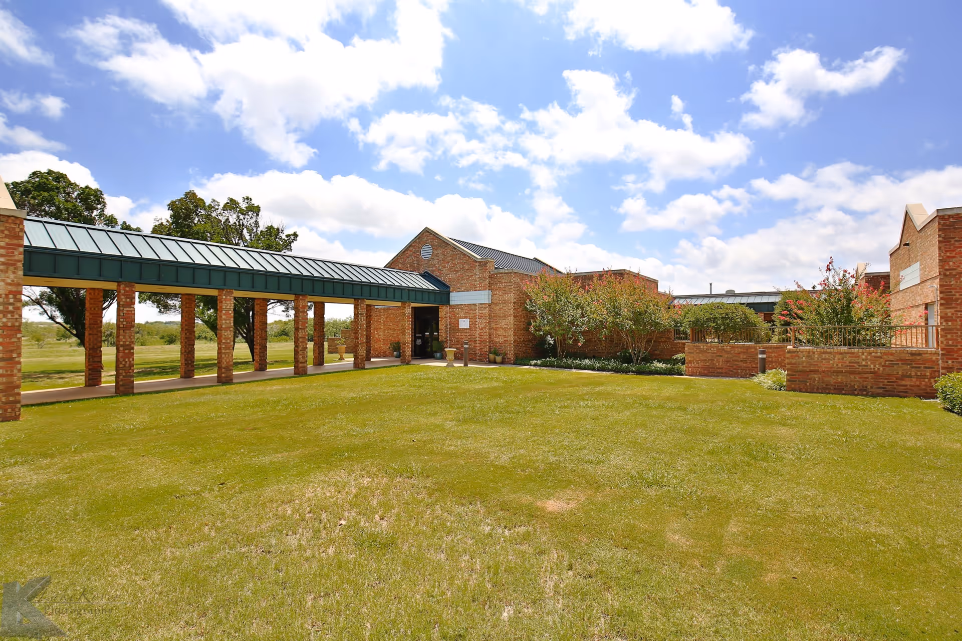 Exterior view of Mesa Springs Healthcare Center showing a brick building with a covered walkway supported by brick columns, surrounded by green grass and trees under a partly cloudy blue sky.