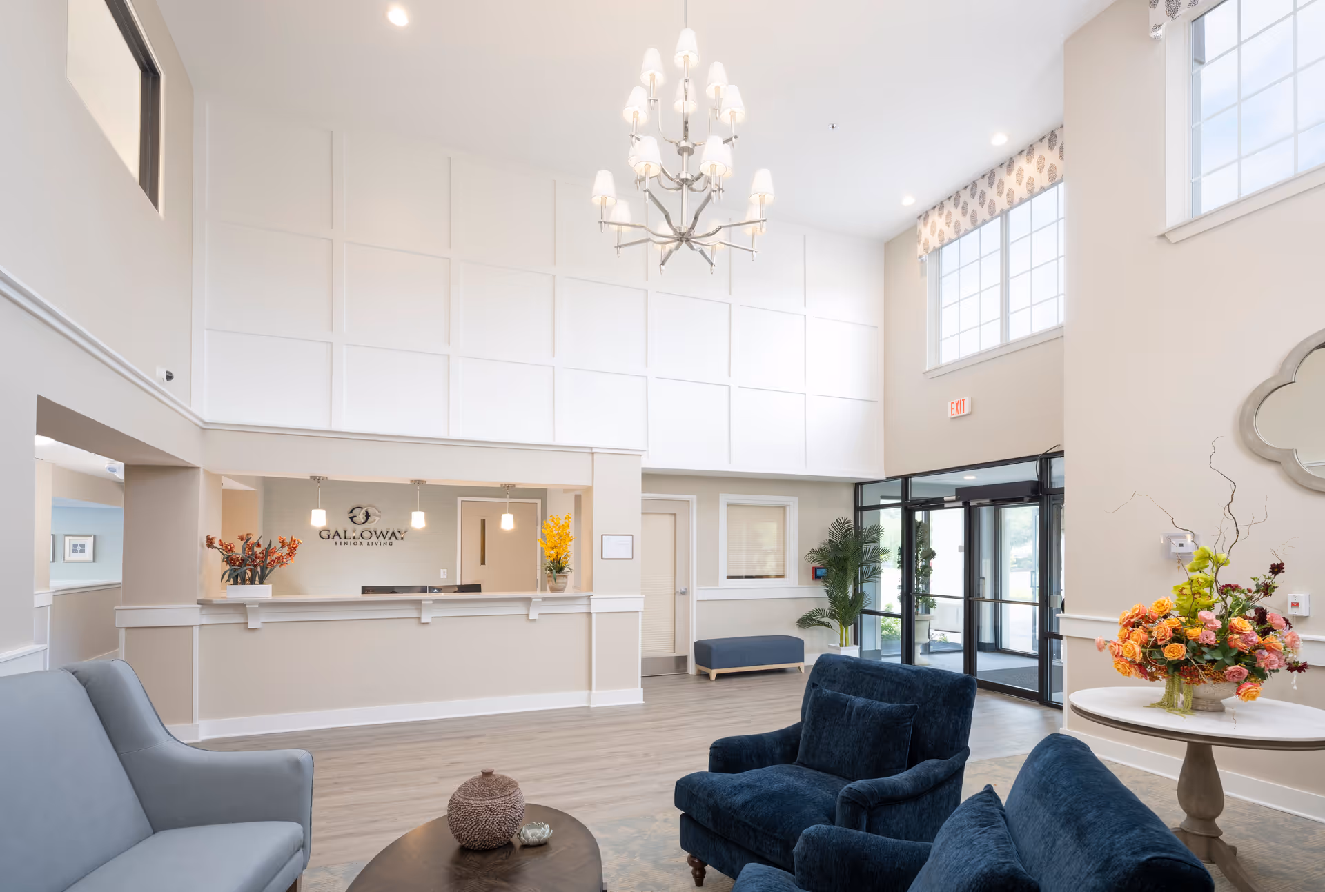 Bright and spacious senior living facility lobby with high ceilings, a chandelier, a reception desk with the Galloway Senior Living logo, comfortable blue armchairs, a gray sofa, a round table with a colorful flower arrangement, large windows, and glass entrance doors.